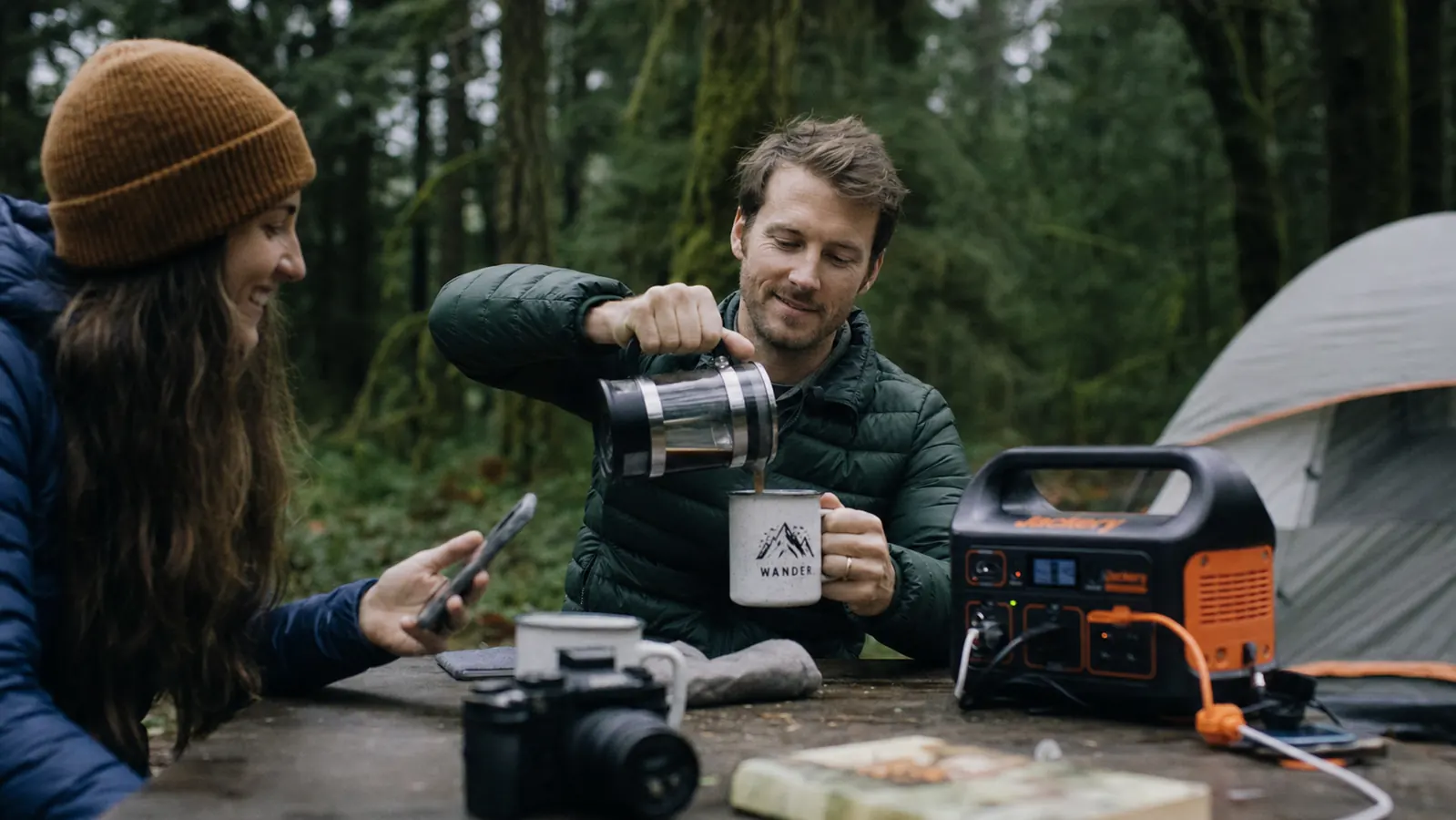 Two campers at a campsite in the morning, one checking a phone while the other pours coffee, a portable power station visible on the table beside their gear